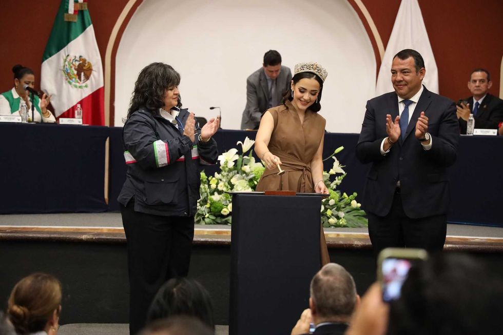 La reina de las fiestas San Juan 2026 cancela el sello postal conmemorativo del 495 aniversario en el Foro San Juan, flanqueada por una representante de Correos de M\u00e9xico y el presidente municipal Roberto Carlos Cabrera Valencia