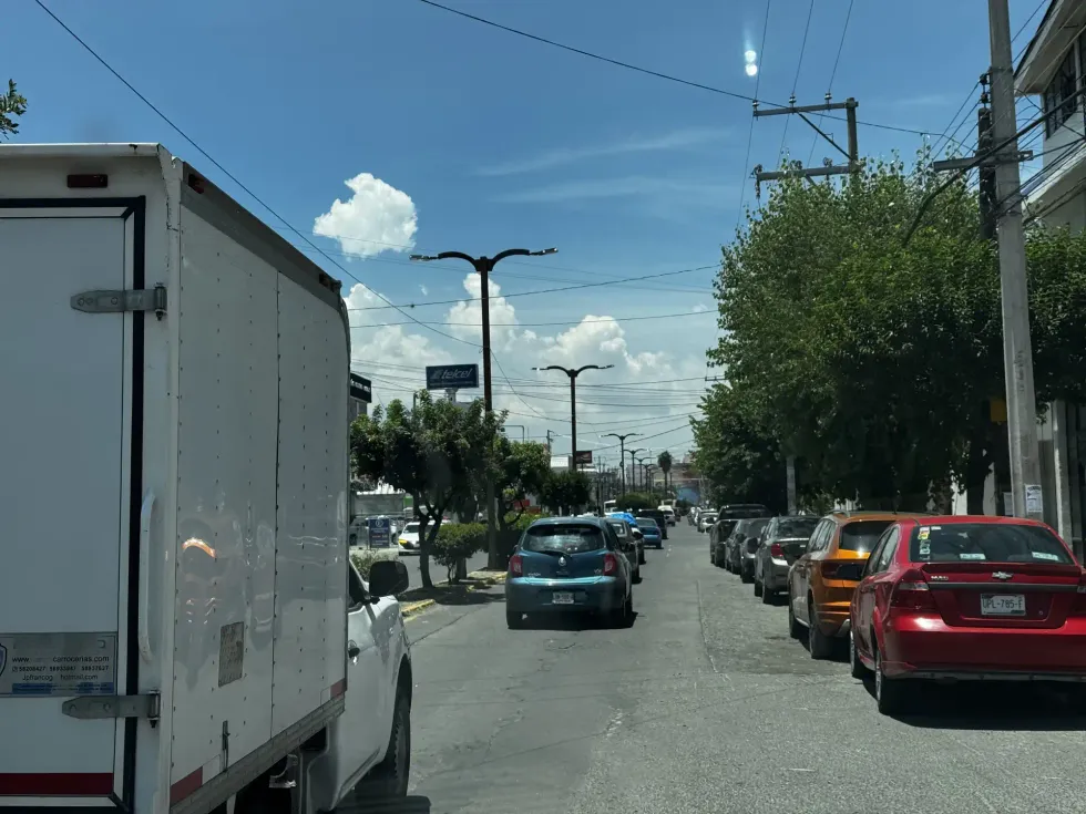 Larga fila de veh\u00edculos detenidos sobre calle adoquinada arbolada en San Juan del R\u00edo, con camioneta de reparto de T\u00eda Rosa en primer plano