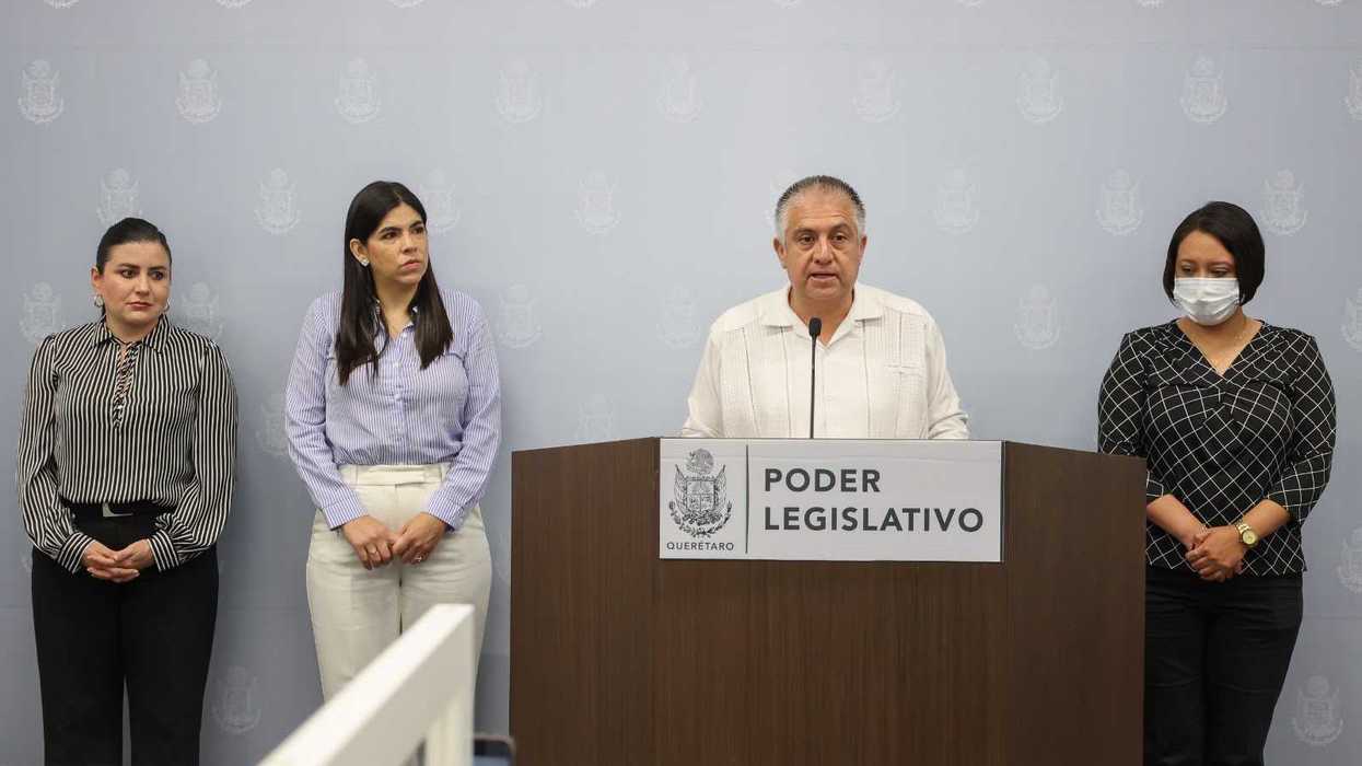 Legislatura de Querétaro: un hombre habla desde el podio del Poder Legislativo mientras tres mujeres permanecen de pie a los lados, frente a un muro con escudos institucionales