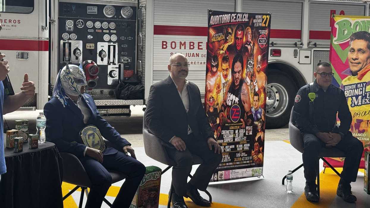 Luchador enmascarado con cinturón de campeón, sentado junto a otros participantes de la presentación y frente al cartel de la función del 27 de noviembre.