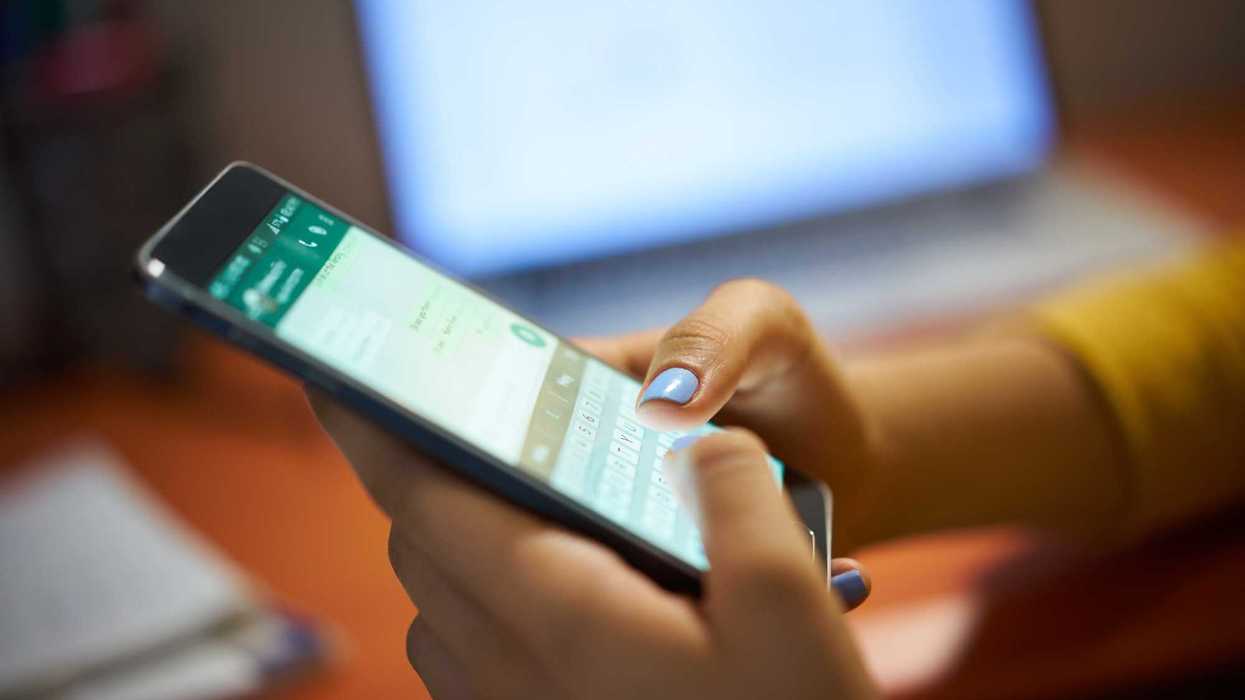 Manos con uñas pintadas de azul escriben en un teléfono con una conversación de mensajería abierta, frente a una computadora portátil, en una imagen de referencia sobre la estafa en WhatsApp con fotos de visualización única.