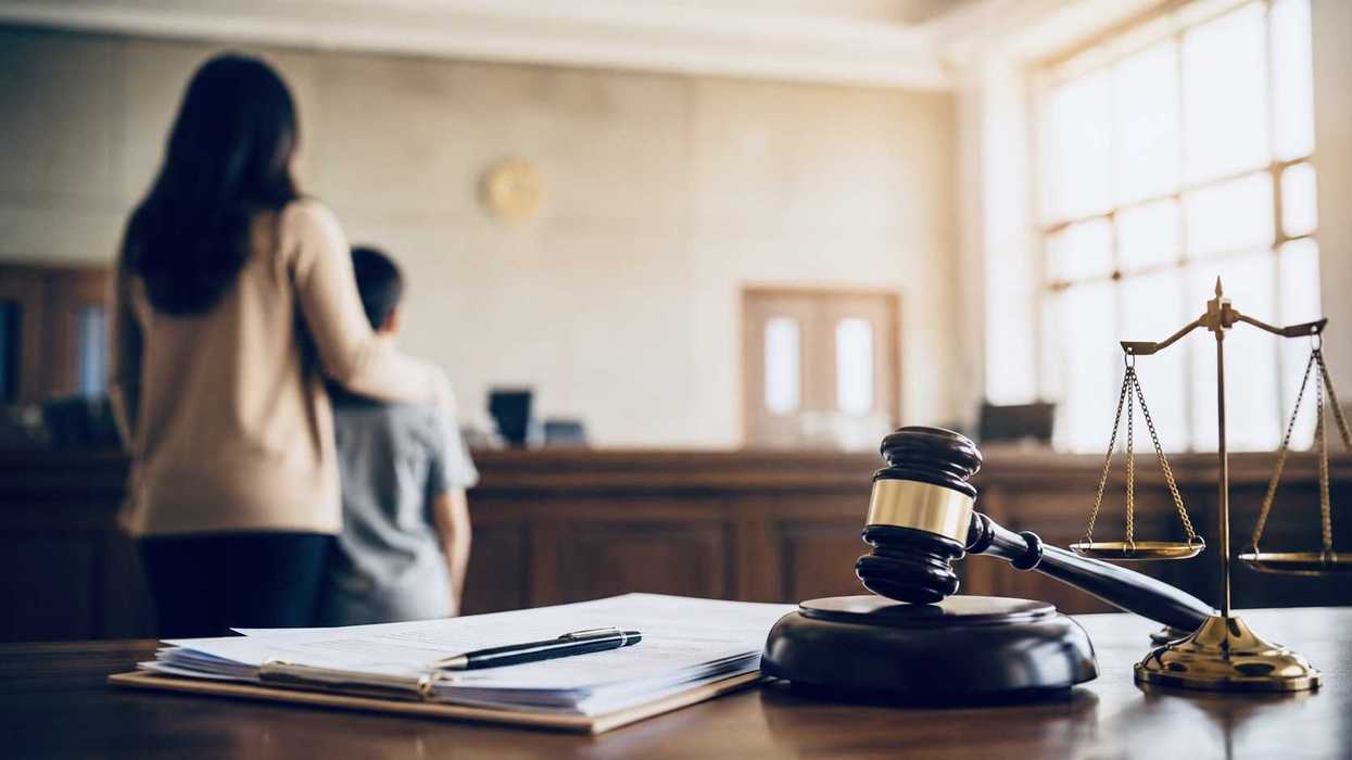 Mazo de juez y balanza de la justicia sobre un escritorio, con una mujer y un niño de espaldas al fondo de una sala.
