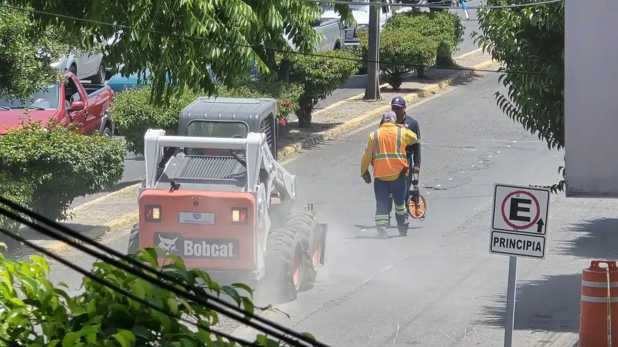 Miniexcavadora Bobcat en movimiento con trabajador con chaleco vial durante labores de bacheo en calle de San Juan del Río.
