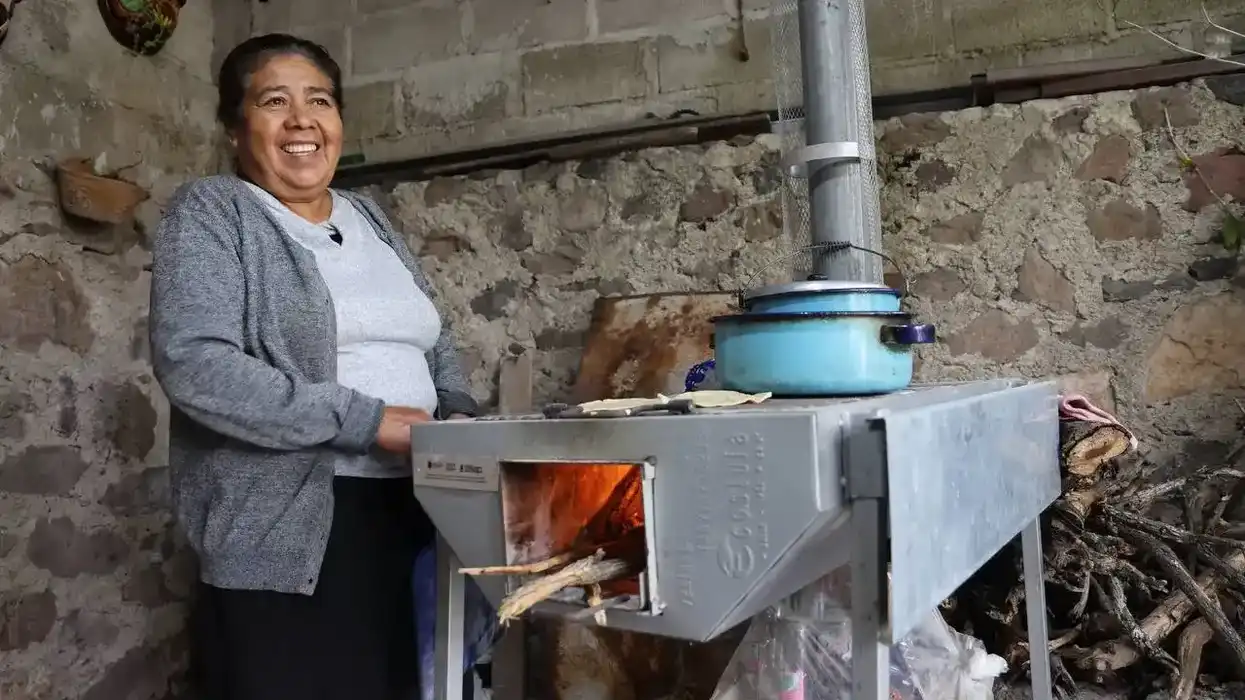 Mujer adulta mayor sonríe junto a su estufa ahorradora de leña encendida, con tortillas sobre la superficie y olla al fuego, en cocina con techumbre rehabilitada. Mesa de Ramírez, Tolimán, Querétaro.