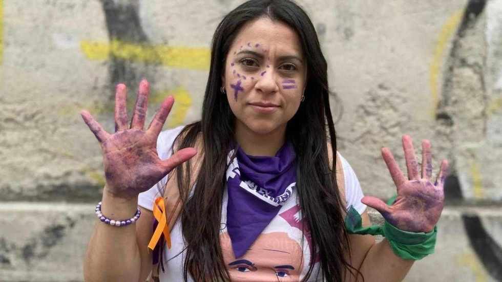 Mujer con las manos pintadas de morado, pa\u00f1uelo morado al cuello y marcas moradas en el rostro, frente a un muro con grafitis