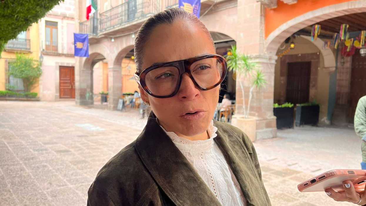 Mujer con lentes y saco verde habla ante celulares de reporteros en una plaza con portales de cantera en Querétaro