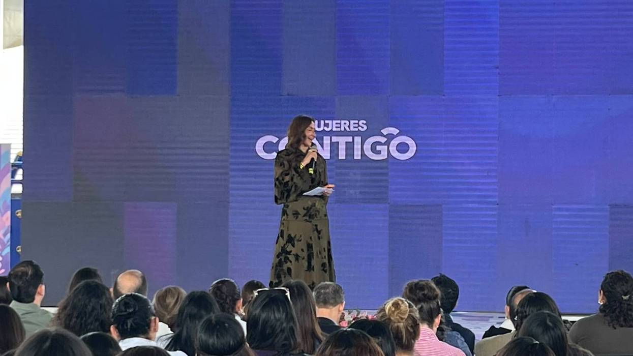 Mujer con micrófono en el escenario de la feria de autonomía económica de mujeres, con el letrero Mujeres Contigo al fondo