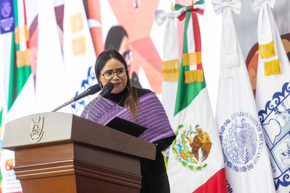 Mujer con rebozo morado habla en el podio del coloquio, junto a banderas
