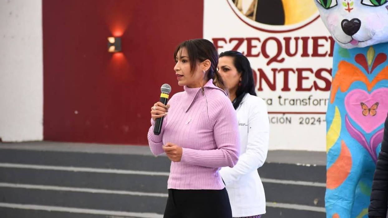 Mujer con suéter rosa habla al micrófono frente a la rotulación del municipio de Ezequiel Montes, junto a una figura de cartonería de colores.
