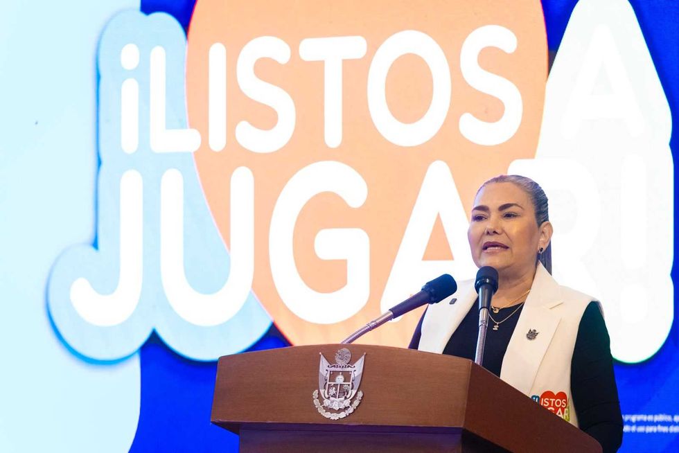 Mujer de chaleco blanco habla en un podio con escudo, frente a la pantalla de Listos a Jugar y con el p\u00fablico en primer plano