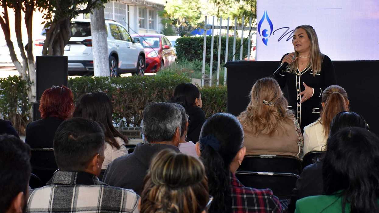 Mujer habla con micrófono ante grupo de asistentes sentados en sesión al aire libre, con pantalla que muestra el logo de la USEBEQ al fondo