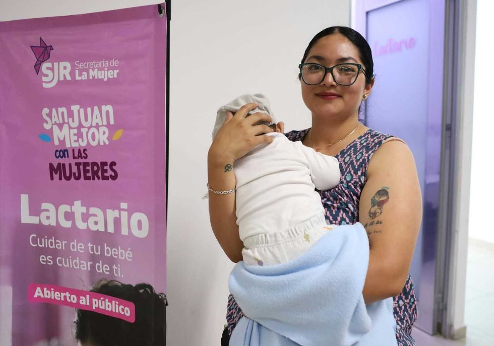Mujer sonr\u00ede con un beb\u00e9 en brazos junto a un cartel del lactario de la Secretar\u00eda de la Mujer.