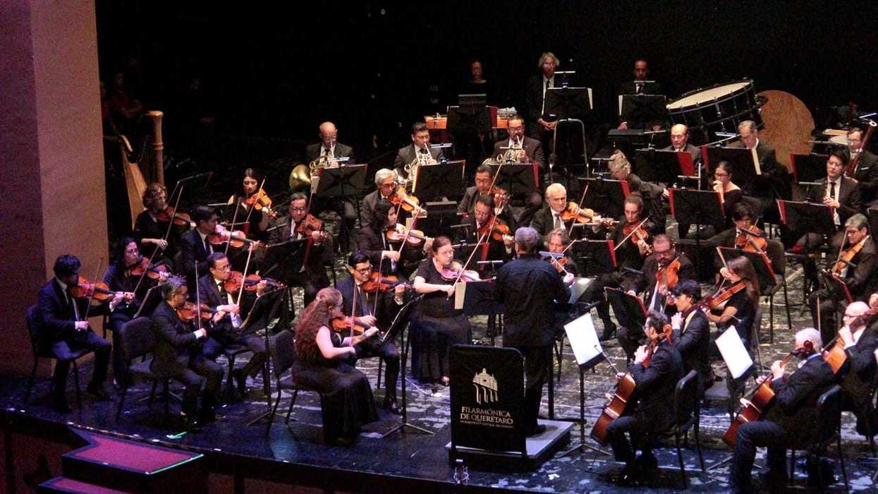 Músicos de la Orquesta Filarmónica de Querétaro en concierto, con las cuerdas al frente, vientos y percusión al fondo y el podio con el nombre de la agrupación.