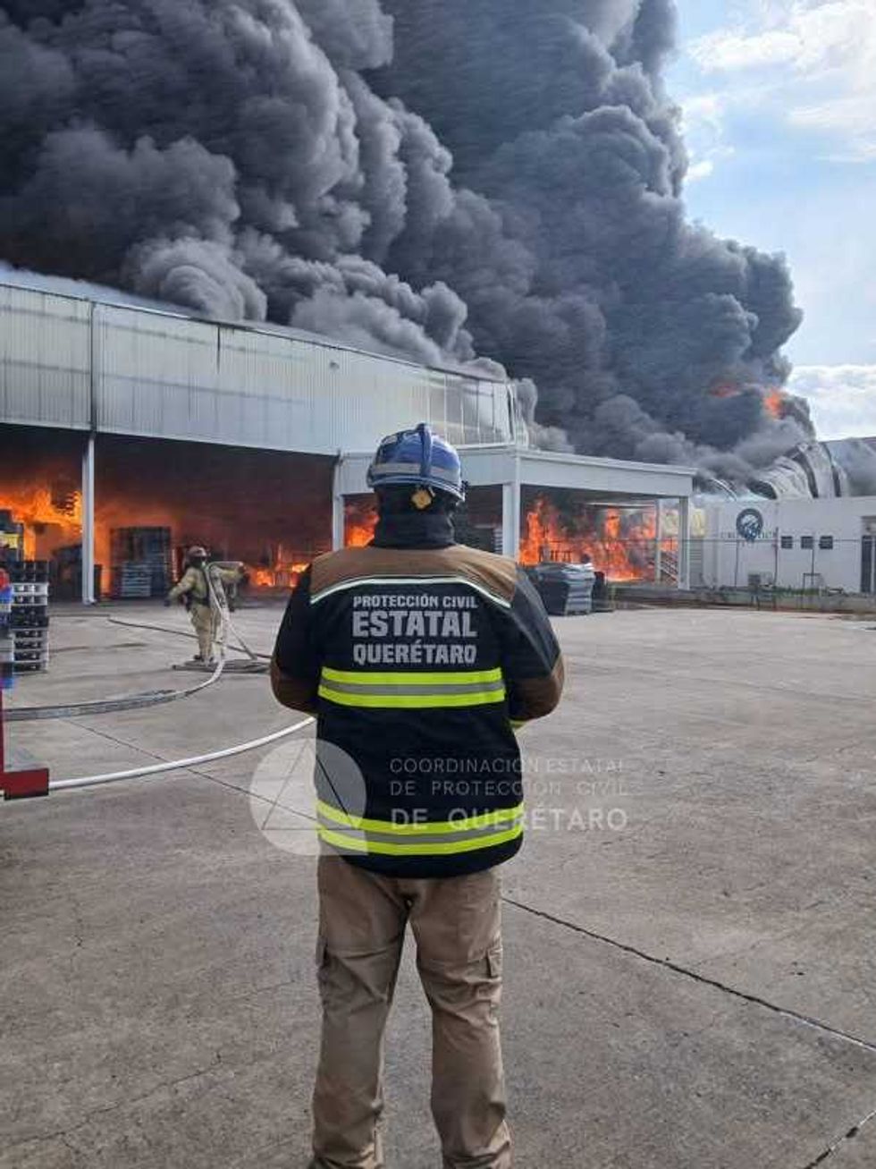 Nave industrial cubierta por una extensa columna de humo negro mientras unidades de emergencia permanecen en el exterior.