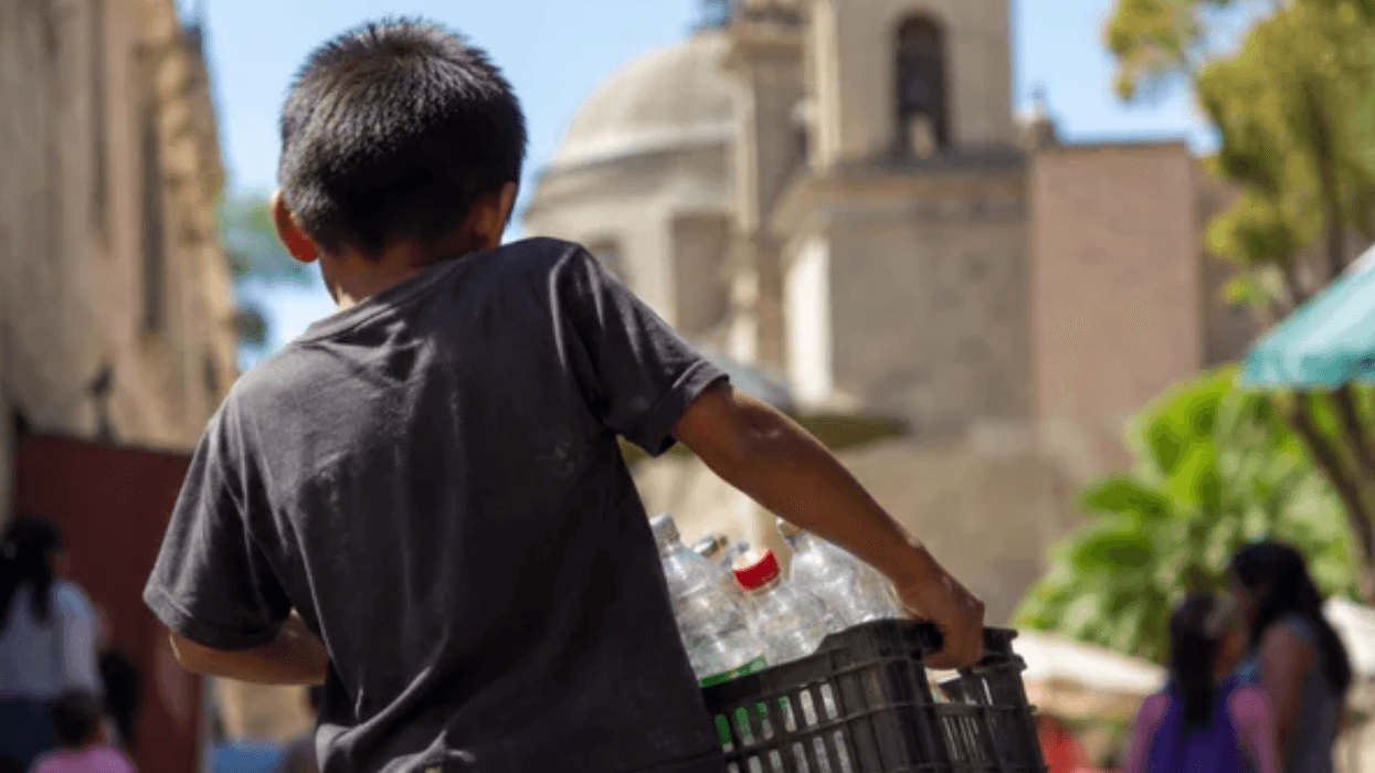 Niño de espaldas carga una caja de plástico con botellas vacías en una calle con un templo al fondo, imagen sobre trabajo infantil en México en 2025.