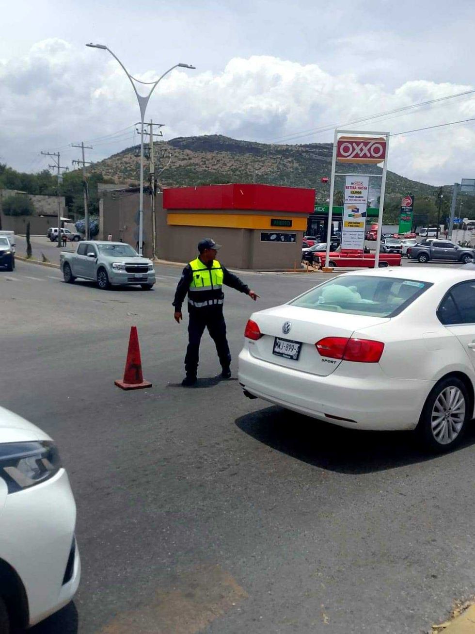 Oficial de tr\u00e1nsito con chaleco reflejante permanece en un cruce peatonal, con una tienda de conveniencia y una gasolinera de fondo y veh\u00edculos, incluida una unidad de transporte p\u00fablico, circulando en la vialidad.