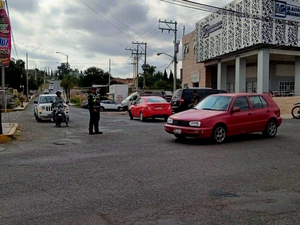 Oficial de tr\u00e1nsito dirige la circulaci\u00f3n vehicular en un cruce con flujo de autom\u00f3viles y una motocicleta, en una zona con edificios comerciales.