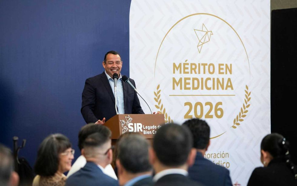 Orador en el atril de la ceremonia del M\u00e9rito en Medicina 2026 en San Juan del R\u00edo, junto al emblema del distintivo