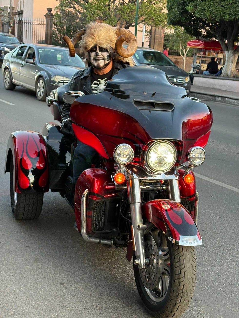 Participante con el rostro pintado de calavera y sombrero de piel con cuernos de carnero sobre un triciclo rojo y negro
