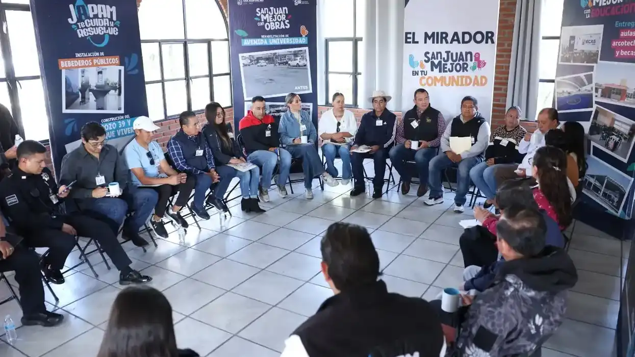 Participantes sentados en círculo en un salón con banners de los programas JAPAM, San Juan Mejor con Obras y El Mirador durante la jornada comunitaria