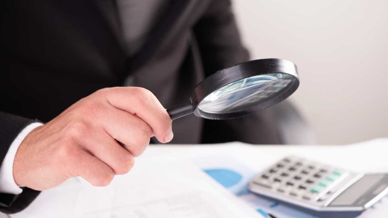 Persona de traje sostiene una lupa sobre documentos junto a una calculadora; imagen ilustrativa de revisión de fondos