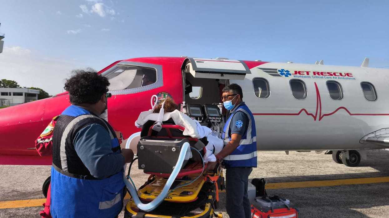 Personal médico y paramédico trasladan en camilla a una paciente hacia una ambulancia, junto a un avión con la puerta abierta en la pista aérea.