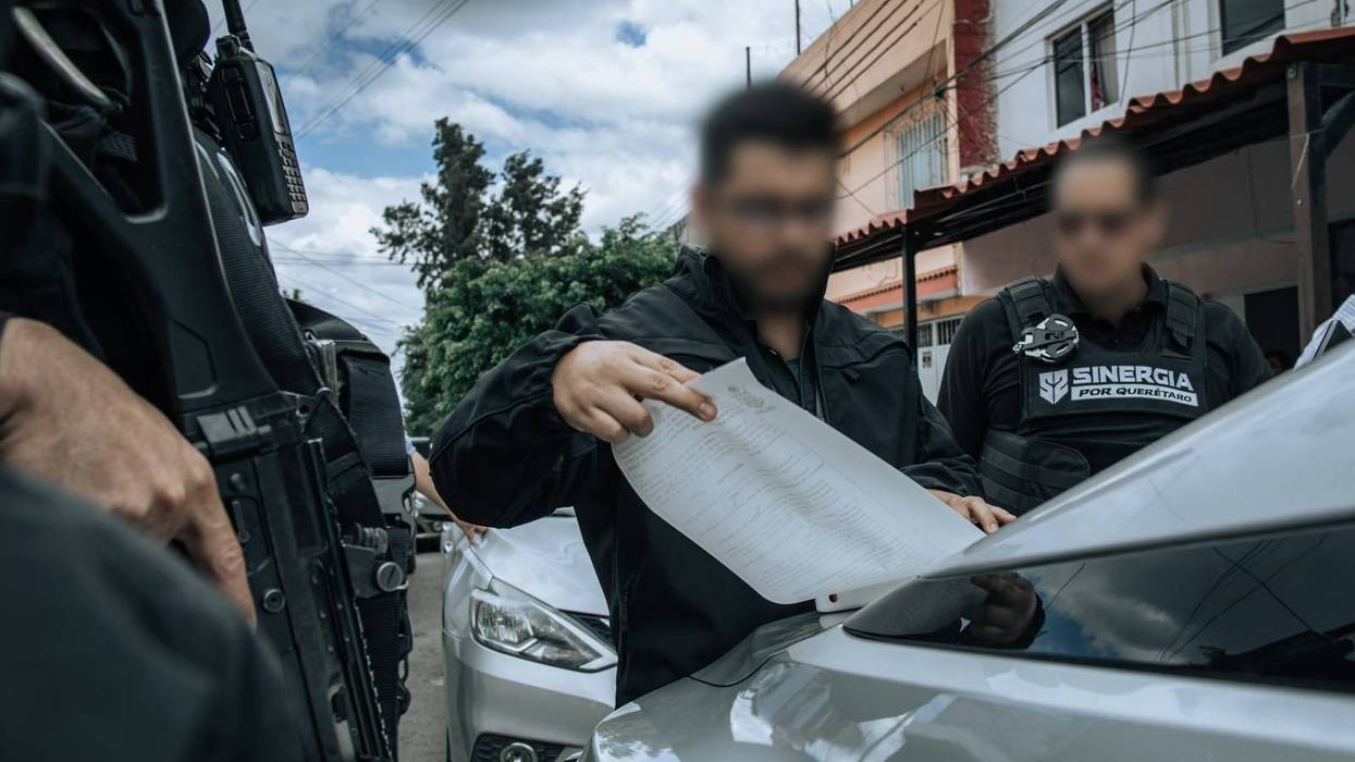 Personal revisa un documento sobre un vehículo junto a un agente armado; Fiscalía de Querétaro investiga homicidios dolosos