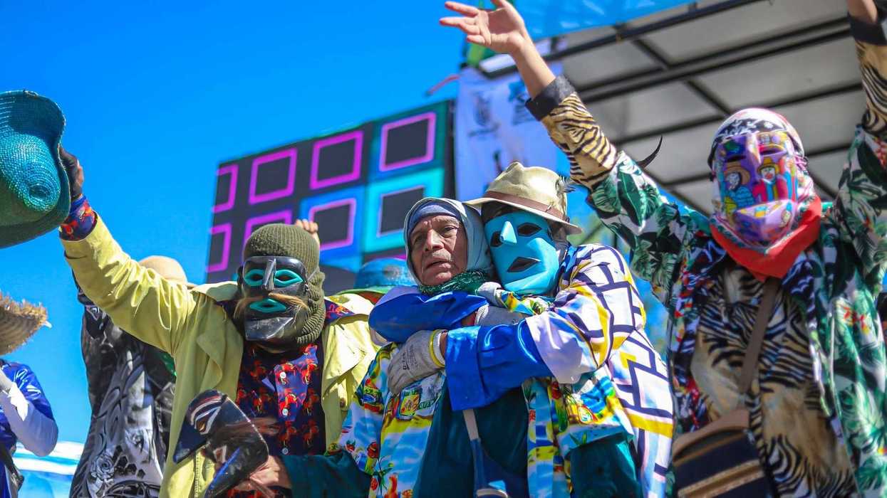 Personas con máscaras de colores y brazos en alto sobre un escenario, una de ellas abraza a un hombre mayor con pañuelo en la cabeza, en un festival de máscaras de La Valla.