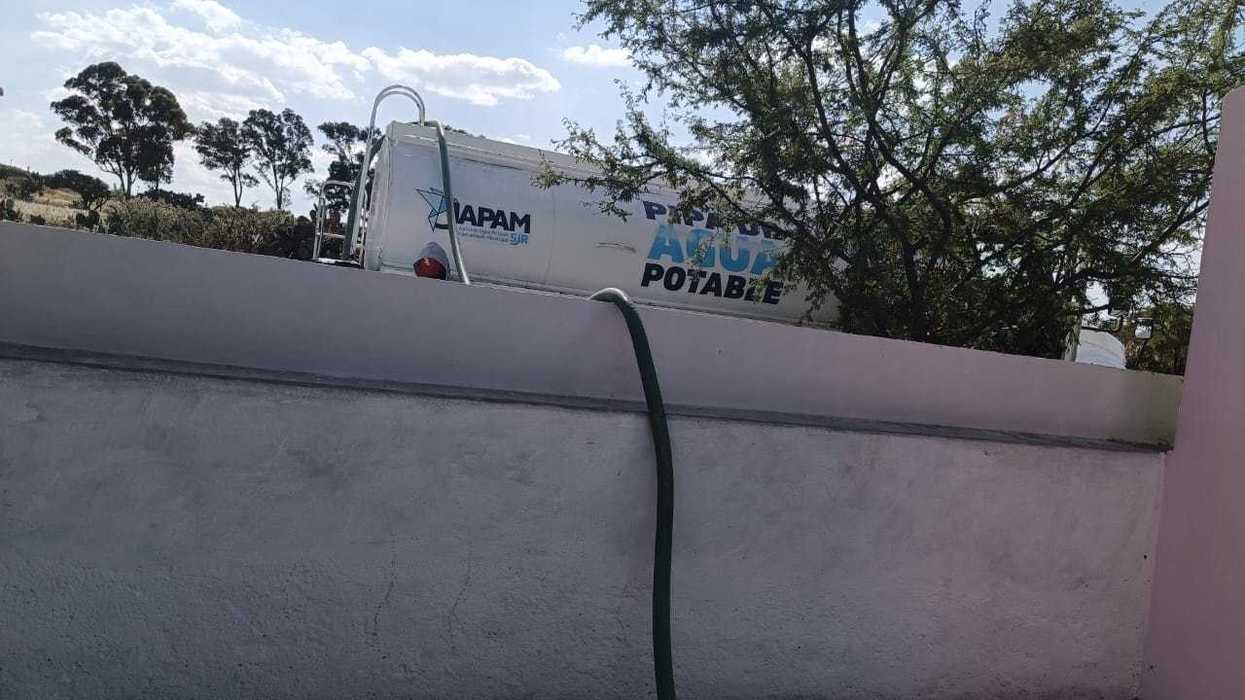 Pipa de agua potable de la JAPAM detrás de una barda, con una manguera verde que cae hacia el exterior, en San Juan del Río