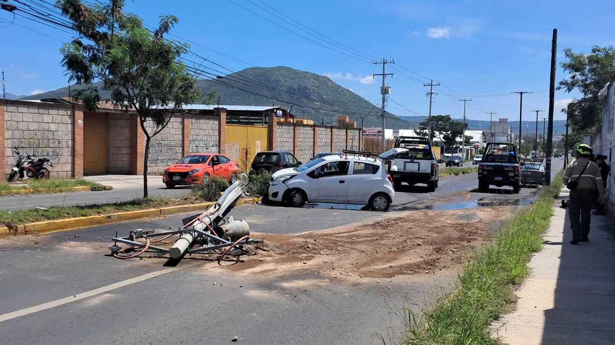 Poste de alumbrado público derribado sobre la vialidad en el sitio del accidente en San Juan del Río, con el vehículo dañado y unidades de policía municipal en segundo plano.