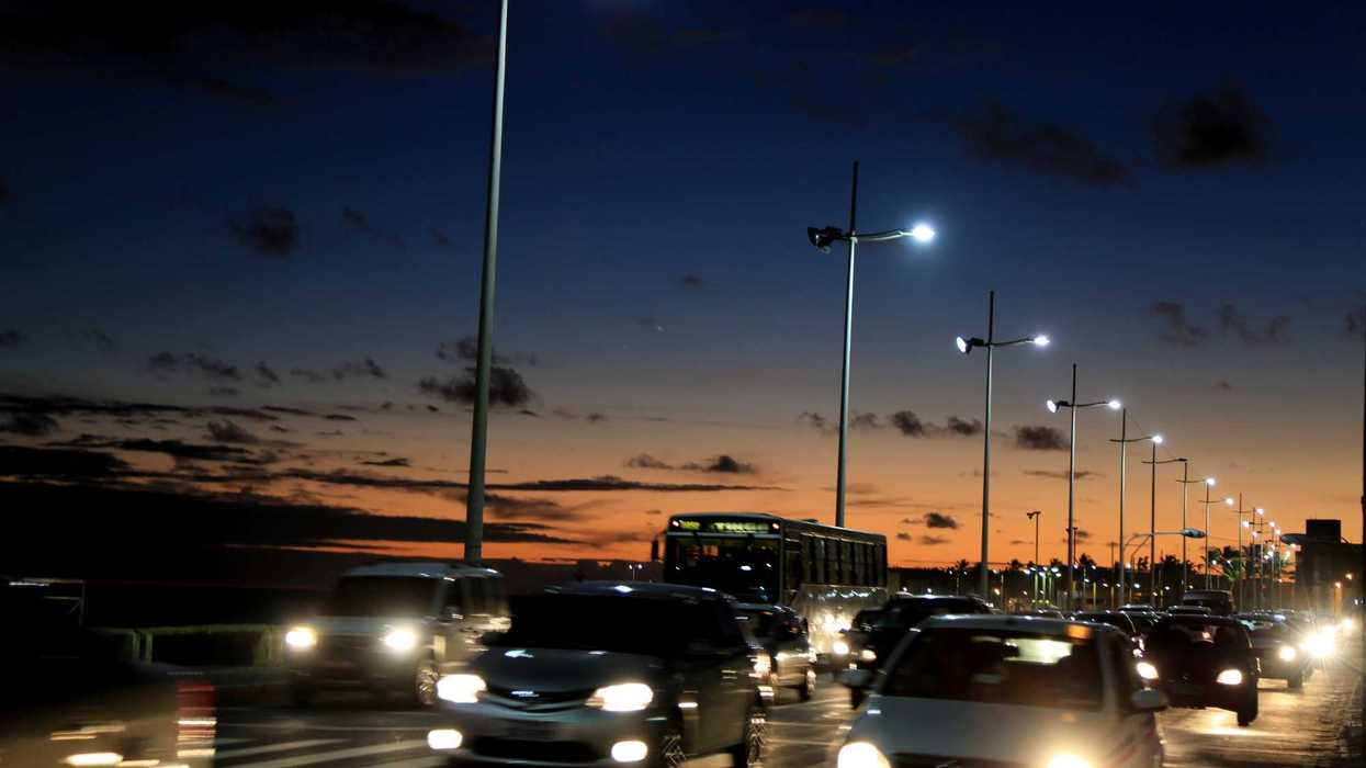 Postes de alumbrado público con luminarias LED encendidas al anochecer sobre una avenida con tránsito vehicular.