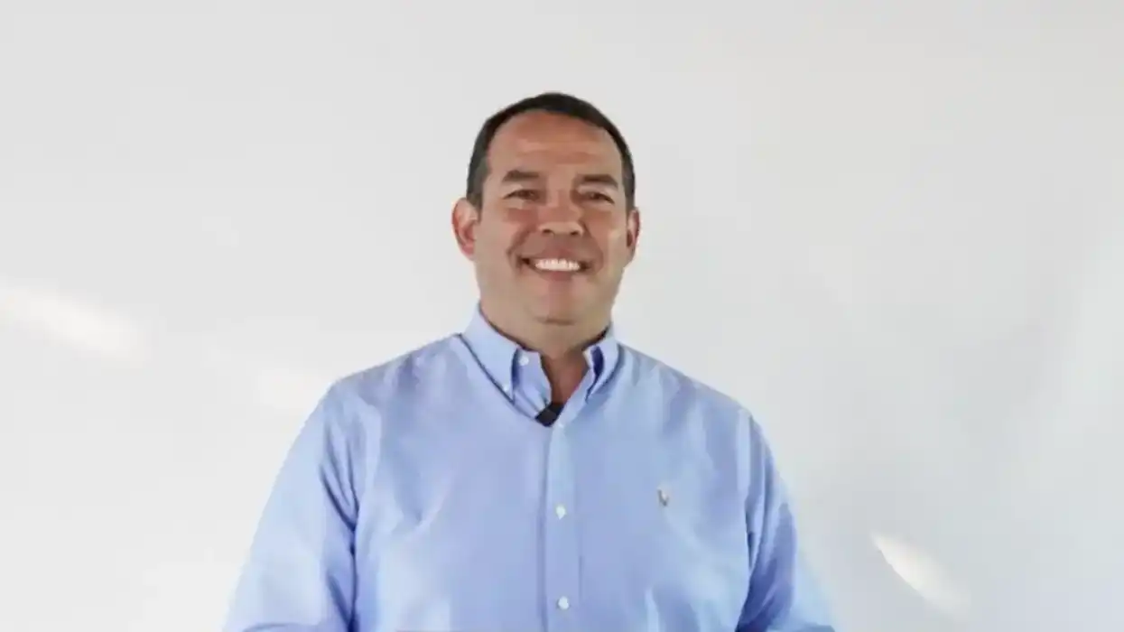 Roberto Cabrera Valencia, alcalde de San Juan del Río, sonríe frente a un podio con camisa azul clara y micrófono de solapa, sobre fondo blanco.