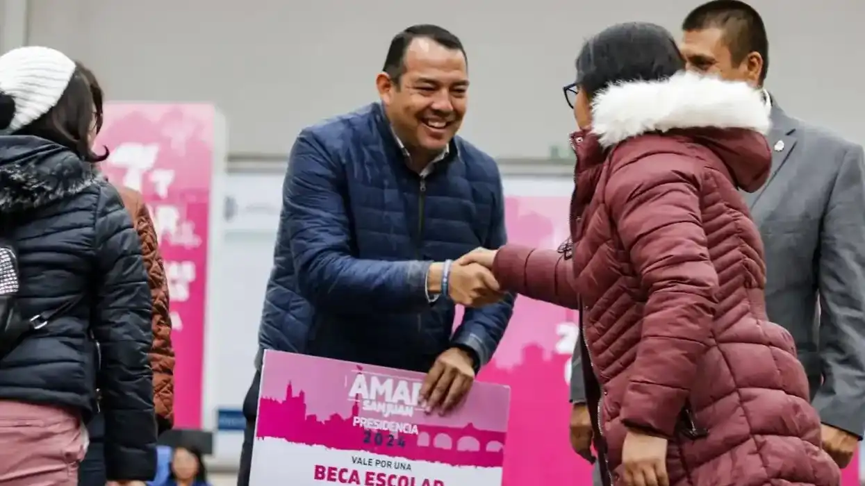 Roberto Cabrera Valencia, alcalde de San Juan del Río, sonríe y estrecha la mano de una mujer mientras sostiene un vale impreso por una beca escolar con la leyenda Amar San Juan Presidencia 2024.