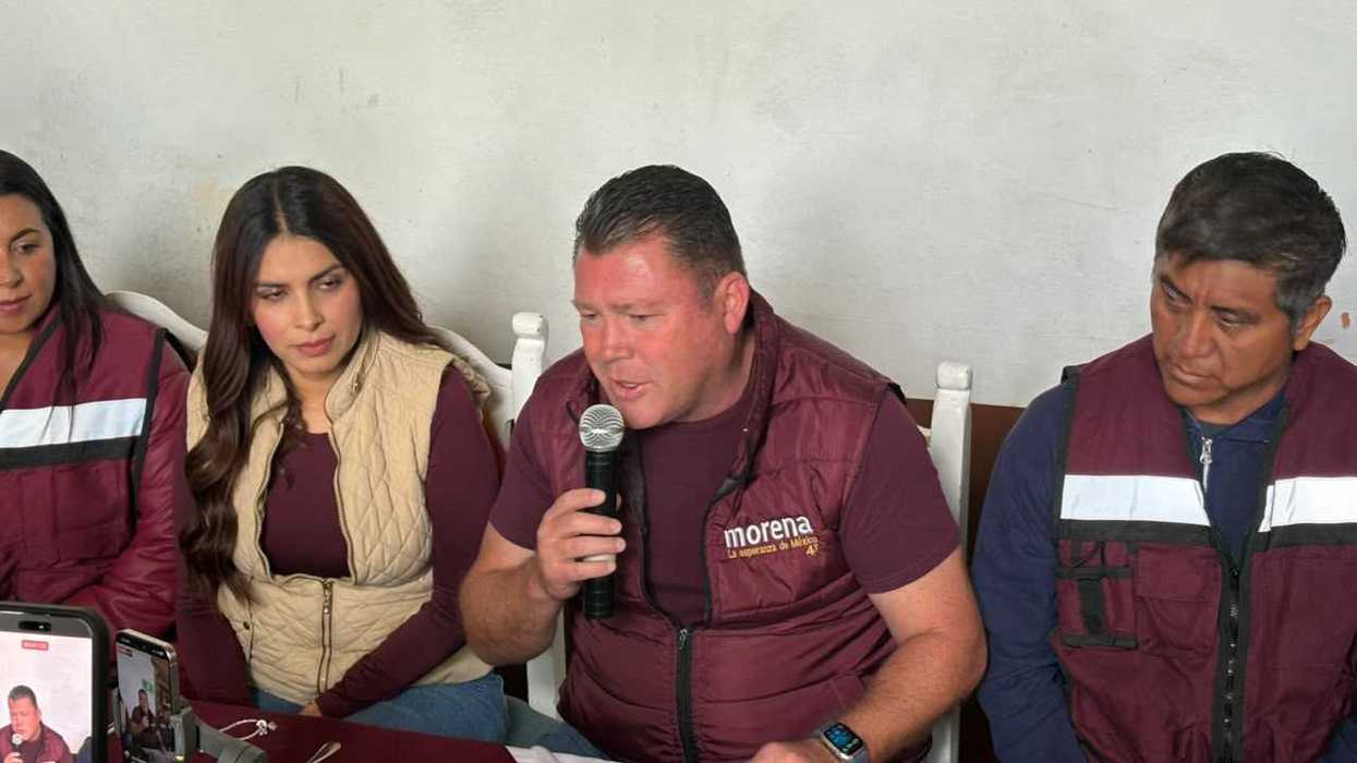 Rueda de prensa de Morena en San Juan del Río: un hombre habla ante el micrófono mientras otras personas con chalecos del partido lo acompañan sentadas.