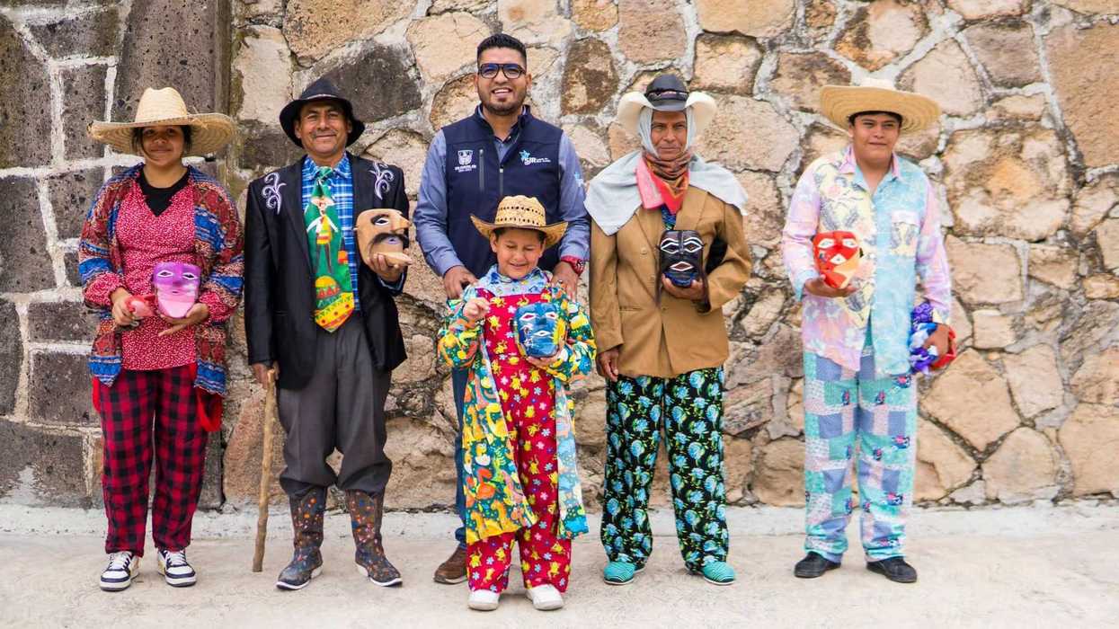 Seis personas posan frente a un muro de piedra, cinco con máscaras de colores en las manos y un hombre con chaleco del municipio de San Juan del Río al centro, en una imagen de los festivales comunitarios de Festivaleando.