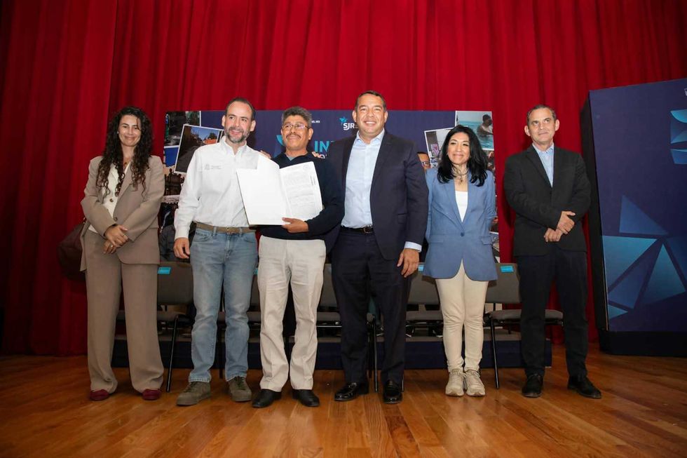 Seis personas posan sobre un escenario con cortina roja y una de ellas sostiene abierta una escritura, en la entrega de escrituras en San Juan del R\u00edo.