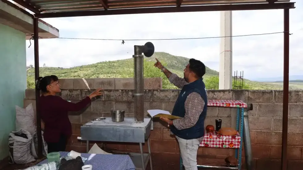 T\u00e9cnico del Sistema Estatal DIF Quer\u00e9taro y mujer beneficiaria de pie bajo techumbre met\u00e1lica rehabilitada, con placa oficial del programa visible, cerro al fondo. Tolim\u00e1n, Quer\u00e9taro.