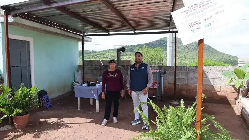 T\u00e9cnico del Sistema Estatal DIF Quer\u00e9taro y mujer beneficiaria de pie bajo techumbre met\u00e1lica rehabilitada, con placa oficial del programa visible, cerro al fondo. Tolim\u00e1n, Quer\u00e9taro.