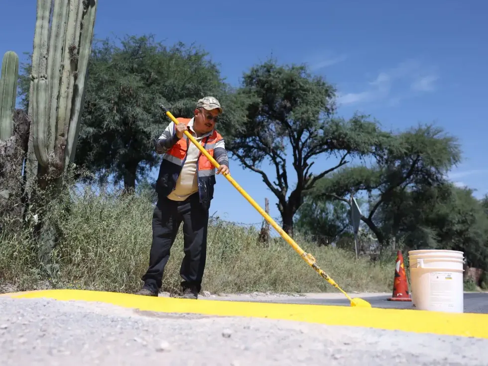 Trabajador con chaleco naranja de Obras P\u00fablicas aplica pintura amarilla en un reductor de velocidad, con cactus y cielo azul al fondo, en la comunidad de El Mirador