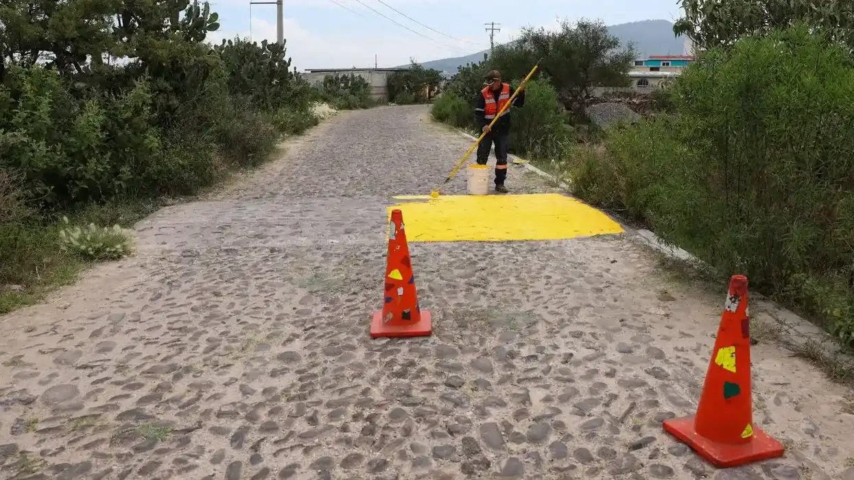 Trabajador con chaleco naranja pinta de amarillo un reductor de velocidad sobre un camino empedrado, entre conos de señalización y vegetación, en la comunidad de Barranca de Cocheros, San Juan del Río.