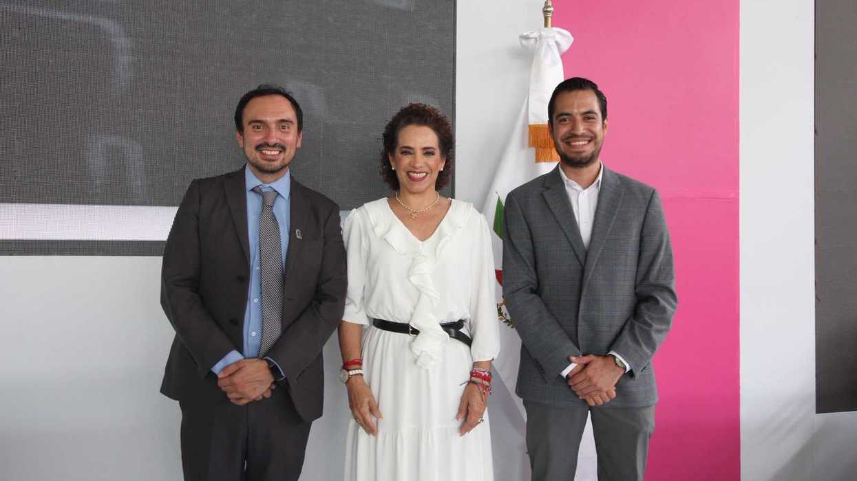Tres personas posan sonrientes frente a una bandera de México y un panel rosa en el evento de la reforma de turismo comunitario en Querétaro.
