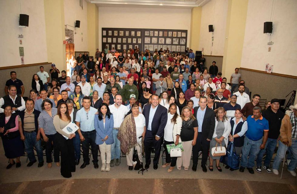 Un grupo de personas posa de pie frente al p\u00fablico sentado en un auditorio durante la entrega de escrituras en San Juan del R\u00edo, una de ellas con una carpeta blanca.