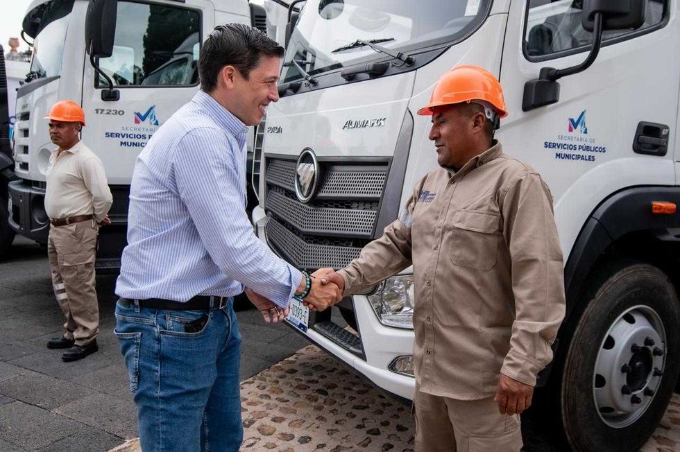 Un hombre con camisa azul a rayas estrecha la mano de un trabajador con uniforme beige y casco naranja junto a un cami\u00f3n de la Secretar\u00eda de Servicios P\u00fablicos Municipales.
