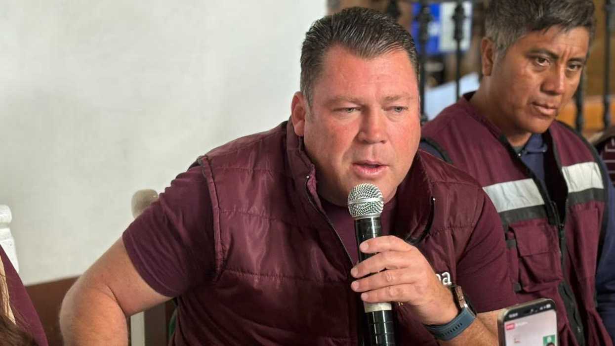 Un hombre con chaleco guinda habla ante el micrófono en una rueda de prensa de Morena sobre la coordinación en San Juan del Río, mientras otra persona escucha detrás.