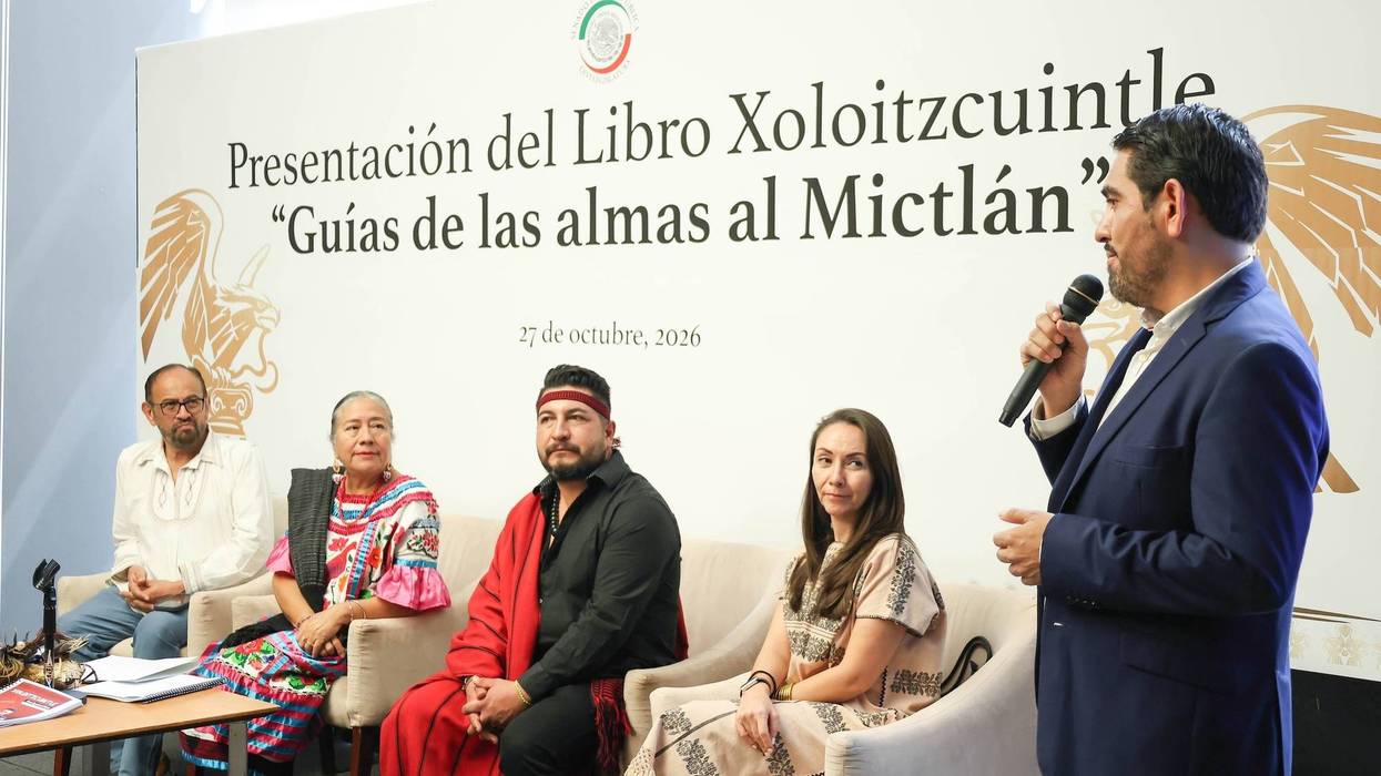 Un hombre habla con micrófono junto a cuatro personas sentadas, ante la manta de presentación del libro sobre el Xolo
