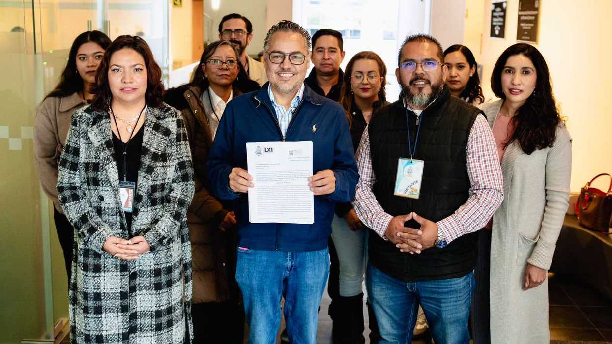 Un hombre sostiene un documento con el logotipo LXI junto a un grupo de personas, varias con gafete de visitante, en un vestíbulo; imagen de la presentación de la iniciativa Escuelas Seguras, Docentes Protegidos en Querétaro.