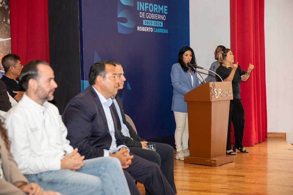 Una mujer con saco azul habla en un podio mientras una int\u00e9rprete de lengua de se\u00f1as interpreta y varios asistentes escuchan sentados en la entrega de escrituras en San Juan del R\u00edo.