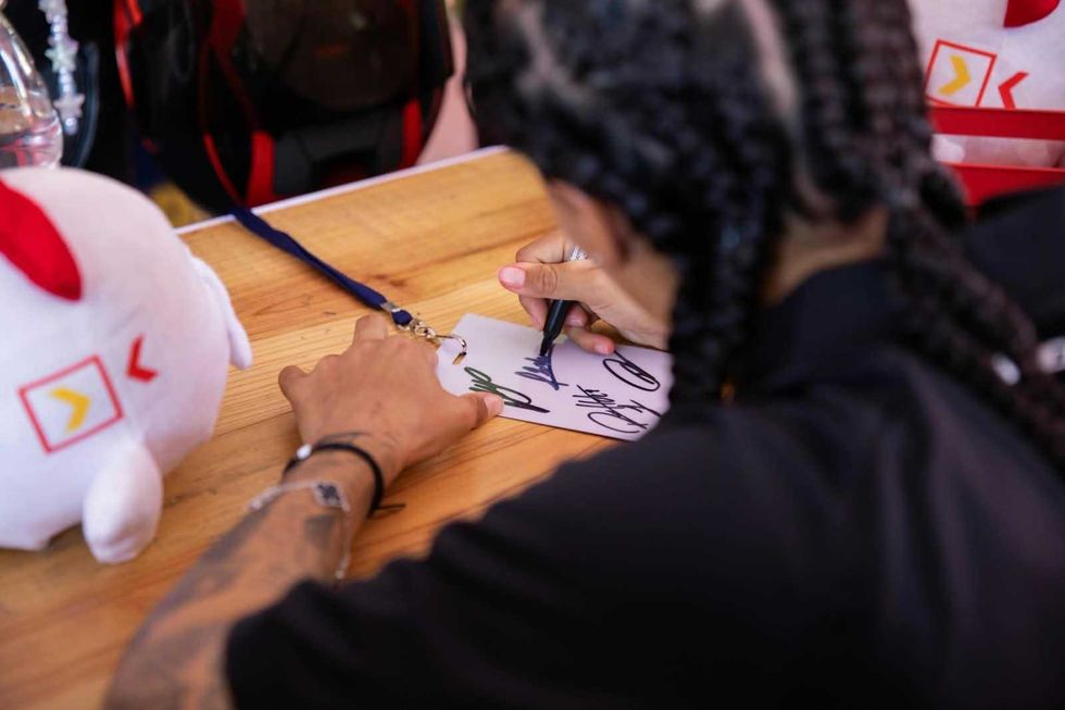 Una persona con trenzas y playera negra firma una l\u00e1mina impresa frente a una lona con la leyenda "Firma de aut\u00f3grafos".