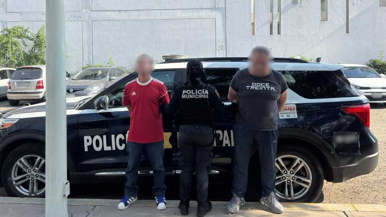 Una policía municipal sujeta a dos hombres de espaldas a una patrulla, con los rostros difuminados.