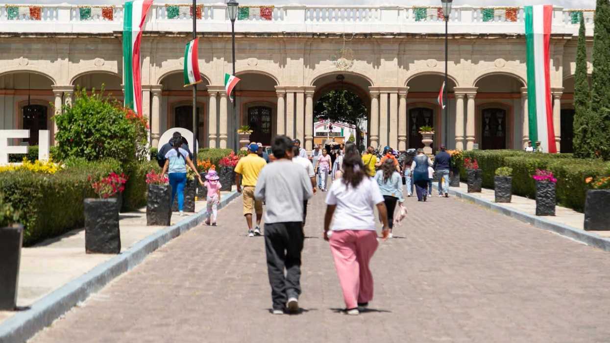Visitantes caminan por un andador empedrado hacia el edificio de arcos de la Ex Hacienda La Llave, con banderas de México en la fachada, durante el Paseo Dominical.